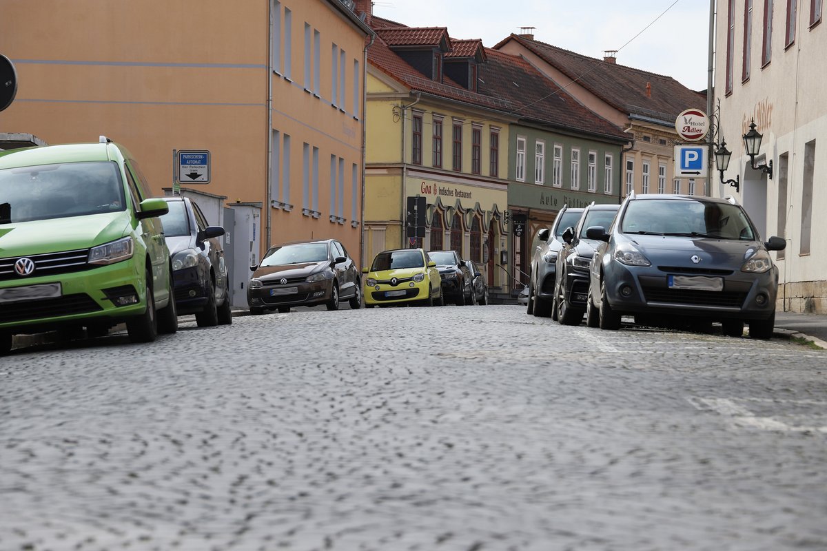 Das Foto zeigt eine Straßenansicht der Mauerstraße in Rudolstadt mit gepflastertem Untergrund und am Straßenrand geparkten Autos unterschiedlicher Marken und Farben. Einige Gebäude mit farblich abgestimmten Fassaden reihen sich entlang der Straße auf, darunter ein Gasthaus und ein Hotel. Im Hintergrund sind weitere historische Gebäude unter einem bewölkten Himmel zu erkennen. Die Szenerie ist menschenleer und vermittelt einen ruhigen Eindruck der städtischen Umgebung.