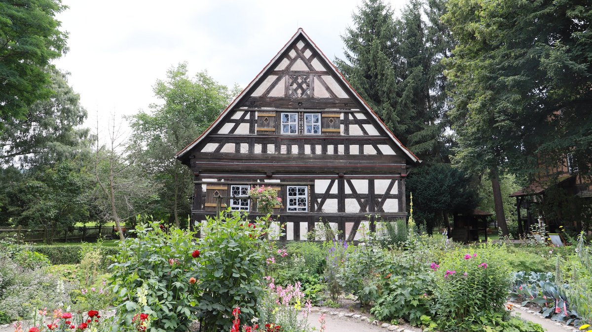 Das Bild zeigt einen gut gepflegten Garten im Vordergrund, umgeben von einem kleinen Zaun und gepflasterten Wegen, die sich durch die Beete schlängeln. Die Beete selbst sind mit einer Vielzahl von Pflanzen, Blumen und Büschen in verschiedenen Höhen und Farben bepflanzt, was eine lebendige und bunte Szenerie ergibt. Im Hintergrund steht ein traditionelles Fachwerkhaus mit sichtbaren Holzbalken und weißen Füllungen dazwischen. Das Dach des Hauses ist steil und mit Ziegeln gedeckt, und die Fenster haben kleine Sprossen. Das Haus wirkt historisch und gut erhalten. Über den Pflanzen und um das Haus herum sind zahlreiche Bäume zu sehen, die zusätzlichen grünen Farbtupfer und Schatten bieten. Das Licht scheint sanft durch die Baumkronen, was auf eine Tageszeit ohne grelle Sonne hinweist, vielleicht Morgen oder Spätnachmittag. Im Hintergrund kann man vereinzelt Menschen erkennen, die scheinbar die Ruhe des Gartens genießen.