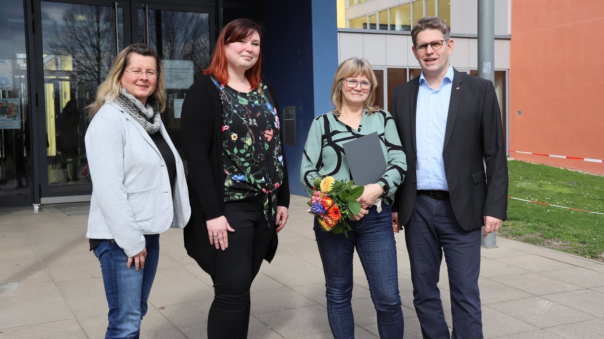 Vier Personen stehen vor dem Eingang eines Schulgebäudes. Von links nach rechts: Eine Frau mit einer hellgrauen Jacke und einem Schal, die lächelnd in die Kamera blickt. Neben ihr steht eine junge Frau mit schwarzer Kleidung und rot gefärbten Haaren, die in die Kamera schaut. Die dritte Person ist eine Frau, die ein grün-gemustertes Top trägt und einen Strauß Blumen sowie eine Mappe in den Händen hält. Sie lächelt und schaut zur Seite. Der vierte im Bunde ist ein Mann in einem dunklen Anzug und einer blauen Krawatte, der in die Kamera lächelt. Sie scheinen gemeinsam einen wichtigen Moment zu teilen, möglicherweise eine Verabschiedung, da die Frau in der Mitte Blumen und eine Mappe hält, was auf eine Ehrung oder ein Abschiedsgeschenk hindeuten könnte. Hinter ihnen ist ein modernes Schulgebäude mit bunten Elementen zu sehen.